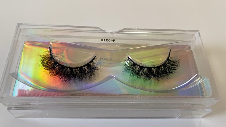 May include: A pair of black false eyelashes in a clear plastic case. The lashes are thick and fluffy, with a natural-looking curl. The case has a holographic insert that reflects a rainbow of colors. The text "M100-F" is printed on the case.