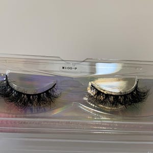 May include: A pair of black false eyelashes in a clear plastic case. The lashes are thick and voluminous, with a curved shape. The case has a holographic iridescent backing. The text "M100-P" is printed on the case.