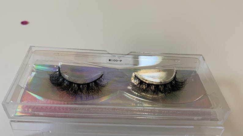 May include: A pair of black false eyelashes in a clear plastic case. The lashes are thick and voluminous, with a curved shape. The case has a holographic iridescent backing. The text "M100-P" is printed on the case.