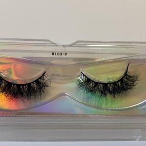 May include: A pair of black false eyelashes in a clear plastic case with a holographic iridescent background. The case is labeled "M100-P". The lashes are thick and fluffy, designed to enhance the eyes.