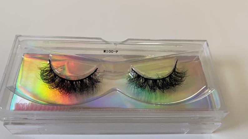 May include: A pair of black false eyelashes in a clear plastic case with a holographic iridescent background. The case is labeled "M100-P". The lashes are thick and fluffy, designed to enhance the eyes.
