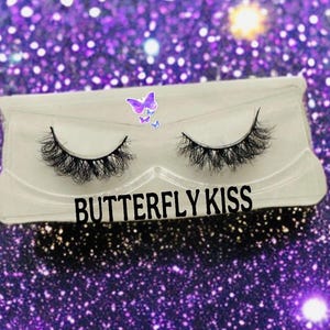 May include: A pair of false eyelashes in a clear plastic case. The case has the words "BUTTERFLY KISS" in black text. A butterfly sticker is on the top left corner of the case. The background is a sparkly purple.
