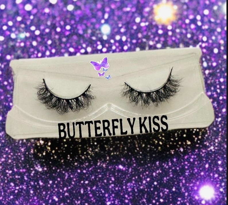 May include: A pair of false eyelashes in a clear plastic case. The case has the words "BUTTERFLY KISS" in black text. A butterfly sticker is on the top left corner of the case. The background is a sparkly purple.
