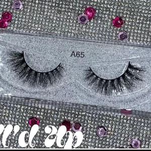 May include: A pair of black false eyelashes in a clear plastic case, labelled "A65". The case sits on a sparkly silver surface scattered with pink and purple gems. The text "Doll'd up" is at the bottom.