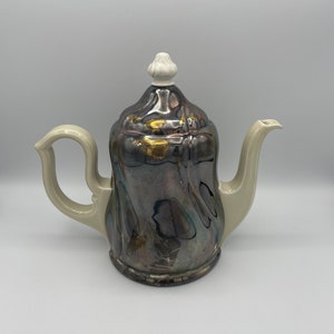 May include: A silver and gold ceramic teapot with a white lid and handle. The teapot has a unique, textured design.