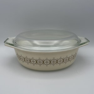 Puede incluir: Una fuente de horno blanca con un patrón geométrico marrón alrededor del borde. La fuente tiene una tapa de vidrio transparente.