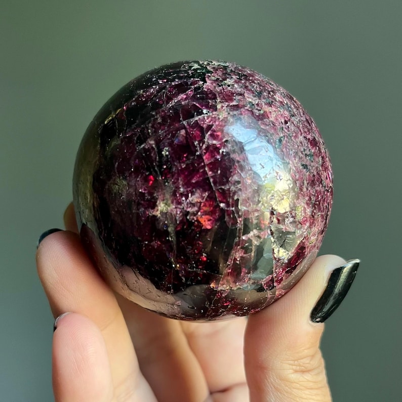 Peut inclure: Une pierre pr&eacute;cieuse sph&eacute;rique polie dans les tons bordeaux fonc&eacute; et marron. La surface de la sph&egrave;re a un aspect craquel&eacute; et textur&eacute; avec des zones tr&egrave;s r&eacute;fl&eacute;chissantes. La pierre est tenue dans une main sur un fond gris doux.
