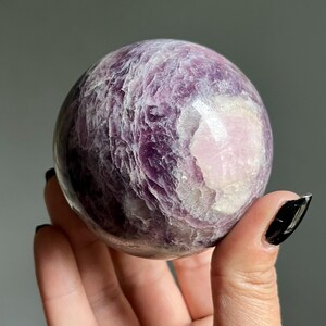 May include: A polished spherical stone featuring a deep purple and lavender color palette with a prominent smooth white patch. The surface displays a layered, crystalline texture with natural variations in opacity and light reflection throughout.