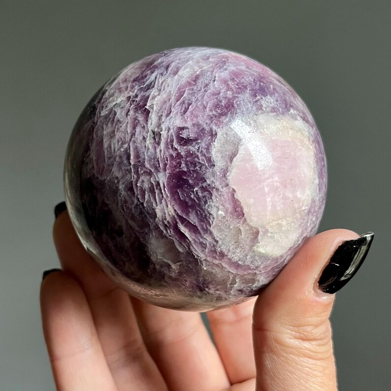 May include: A polished spherical stone featuring a deep purple and lavender color palette with a prominent smooth white patch. The surface displays a layered, crystalline texture with natural variations in opacity and light reflection throughout.