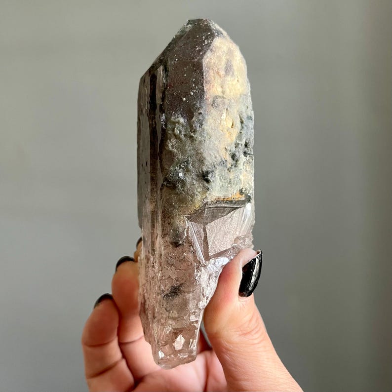 Op de afbeelding: Een natuurlijk kwartskristal met een puntige punt en een ruw, getextureerd oppervlak. Het kristal heeft een kleurengradi&euml;nt, waaronder helder, bruin en beige. Het kristal wordt in een hand gehouden.