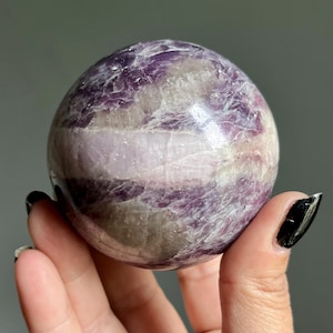 May include: A polished spherical stone featuring a mix of deep purple, lavender, and translucent white mineral bands. The surface displays natural crystalline textures and internal fractures throughout the orb, creating a varied color pattern.