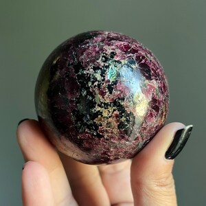 Peut inclure: Une pierre pr&eacute;cieuse sph&eacute;rique polie, aux couleurs bordeaux fonc&eacute; et noir. La surface pr&eacute;sente un aspect textur&eacute; avec des &eacute;clats d'or et de vert iris&eacute;. La sph&egrave;re est tenue dans une main, mettant en valeur sa taille et ses d&eacute;tails.