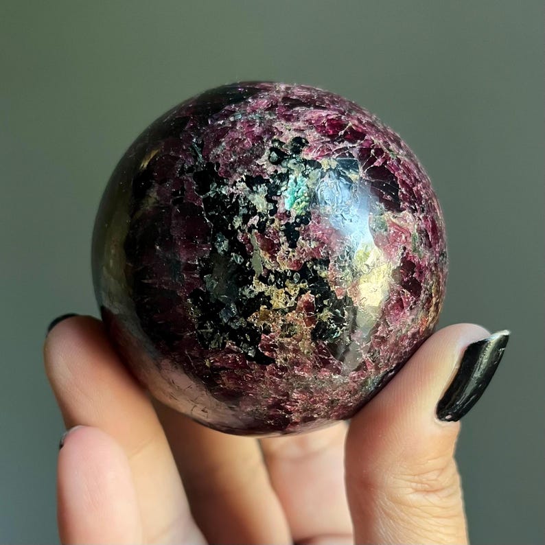 Peut inclure: Une pierre pr&eacute;cieuse sph&eacute;rique polie, aux couleurs bordeaux fonc&eacute; et noir. La surface pr&eacute;sente un aspect textur&eacute; avec des &eacute;clats d'or et de vert iris&eacute;. La sph&egrave;re est tenue dans une main, mettant en valeur sa taille et ses d&eacute;tails.