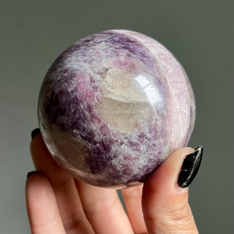 May include: A polished spherical stone featuring a marbled pattern of deep purple, lavender, and translucent white mineral inclusions. The surface is smooth and reflective, showcasing natural crystalline textures and varied color gradients throughout.