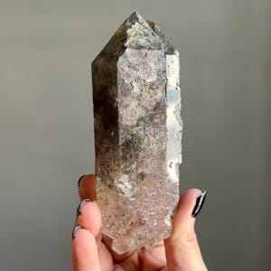Op de afbeelding: Een heldere kwartskristal met een puntige top en een ruw, getextureerd oppervlak. Het kristal heeft een mix van kleuren, waaronder helder, bruin en groen. Het kristal wordt in een hand gehouden.