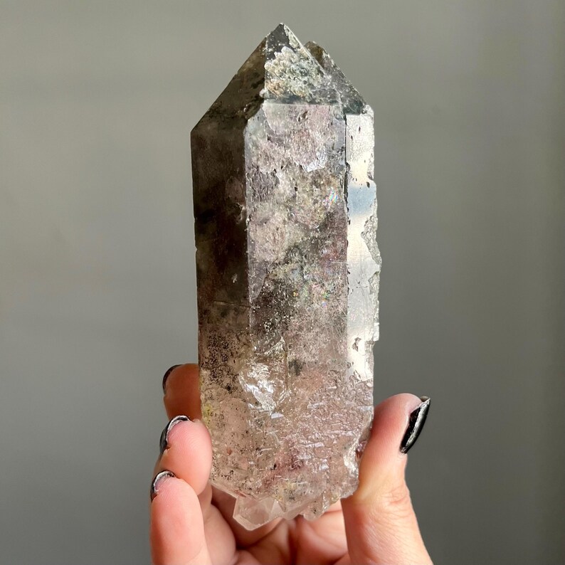 Op de afbeelding: Een heldere kwartskristal met een puntige top en een ruw, getextureerd oppervlak. Het kristal heeft een mix van kleuren, waaronder helder, bruin en groen. Het kristal wordt in een hand gehouden.