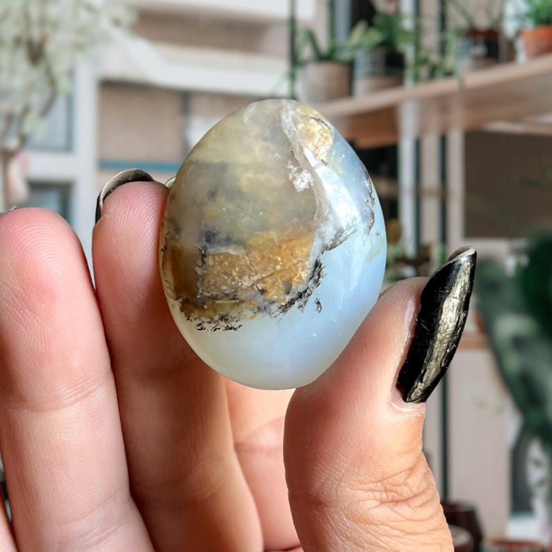 Op de afbeelding: Een gladde, ovale, lichtblauwe en witte opaal edelsteen met bruine en zwarte insluitsels. De edelsteen wordt in een hand gehouden.