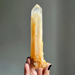 May include: A tall, translucent quartz crystal point with a natural golden-yellow hue. The mineral features internal golden inclusions and a rough, crystalline base with jagged edges, tapering to a smooth, pointed termination at the top.