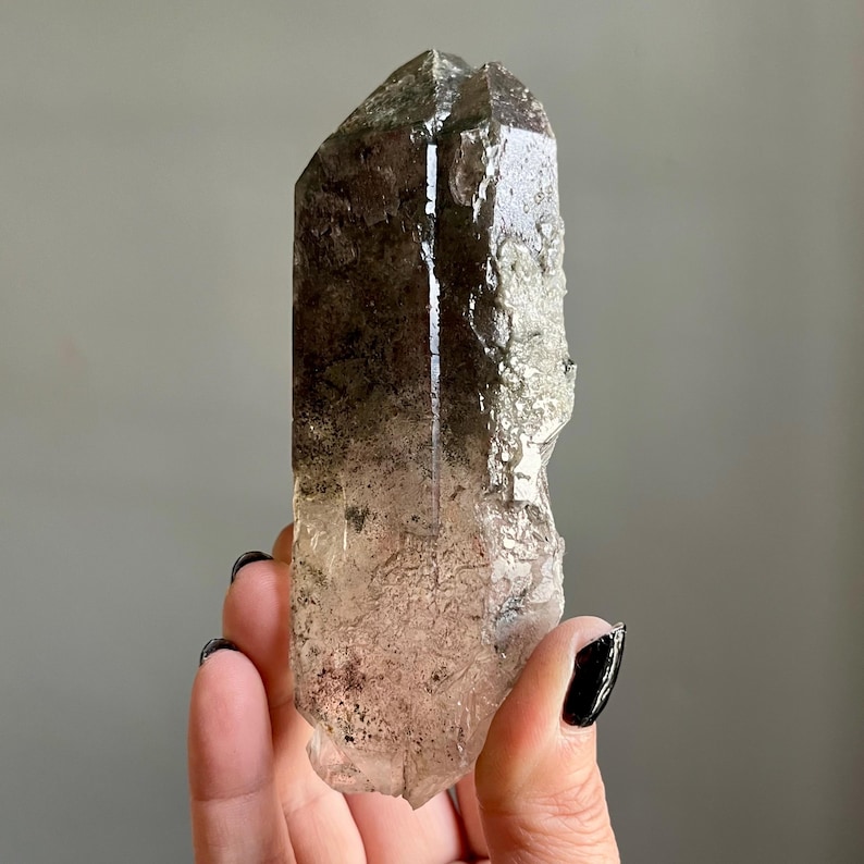 Op de afbeelding: Een natuurlijke rookkwarts kristalpunt. Het kristal heeft een doorschijnende bruine kleur met een ruw, getextureerd oppervlak. De punt wordt in een hand gehouden tegen een grijze achtergrond. Het kristal is een natuurlijke, ruwe edelsteen.
