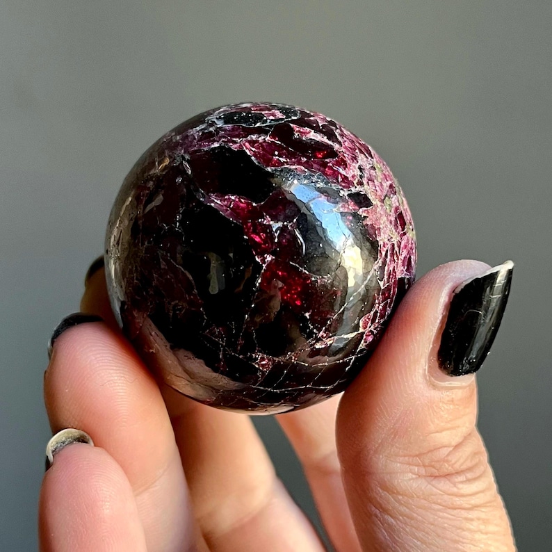 May include: A polished, spherical gemstone with a deep burgundy and black color scheme. The surface has a cracked, textured appearance with veins of lighter red. The sphere is held in a hand, showcasing its size and detail.