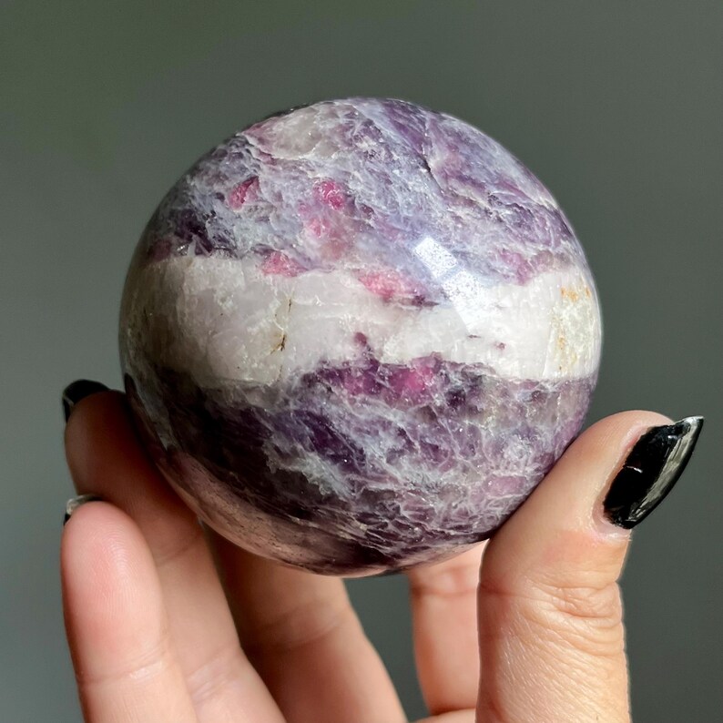 May include: A polished spherical stone featuring a mix of deep purple, magenta, and white mineral bands. The surface displays a natural crystalline texture with varied opacity and reflective highlights across its rounded, smooth, and solid form.