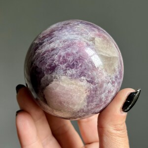 May include: A polished spherical stone featuring a marbled texture with deep purple, lavender, and creamy white mineral inclusions. The surface is smooth and reflective, showcasing natural crystalline patterns throughout the round mineral specimen.