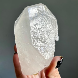 May include: A clear, translucent quartz crystal point featuring a multifaceted, geometric surface texture with triangular etchings and natural striations. The mineral specimen has a pointed termination and a semi-opaque base, displaying internal fractures.