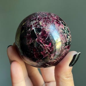 Peut inclure: Une sph&egrave;re de pierre pr&eacute;cieuse polie, de couleur bordeaux fonc&eacute; et noire. La surface pr&eacute;sente un aspect craquel&eacute; et textur&eacute;, avec des veines plus claires. La sph&egrave;re est tenue dans une main, mettant en valeur sa taille et ses d&eacute;tails.