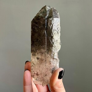 Op de afbeelding: Een natuurlijke rookkwarts kristalpunt. Het kristal heeft een verloop van helder naar donkerbruin, met een ruw, getextureerd oppervlak. De punt is een donkerdere tint bruin. Het kristal wordt door een persoon vastgehouden.