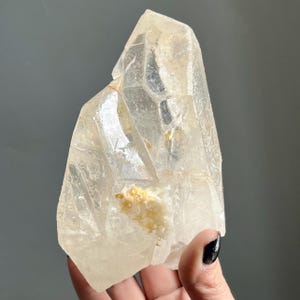 May include: A clear, translucent quartz crystal specimen with a pointed termination and multifaceted surfaces. The mineral features internal fractures and a small, textured patch of opaque white and pale yellow mineral deposits on its front face.