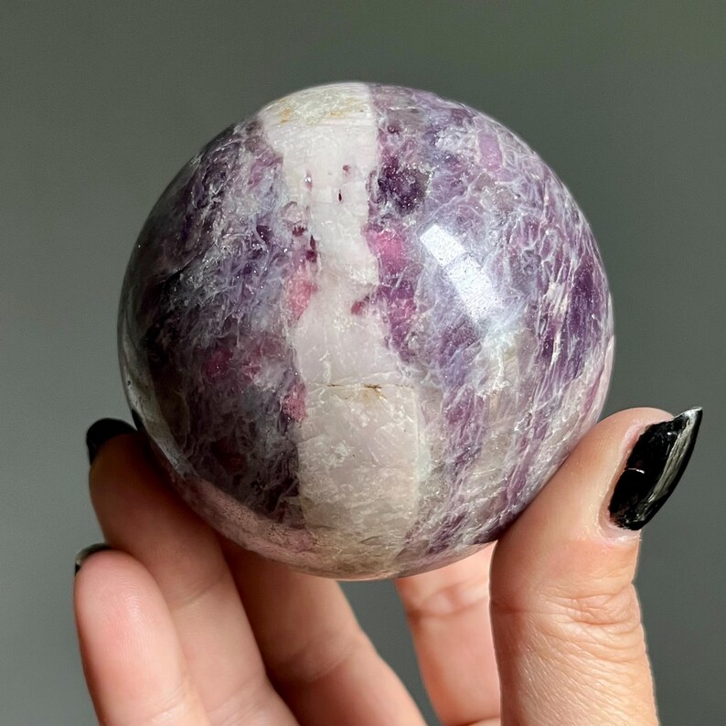 May include: A polished spherical stone featuring a prominent vertical band of opaque white mineral running through a deep purple and magenta crystalline body. The surface displays natural fractures and varied textures within the polished sphere.