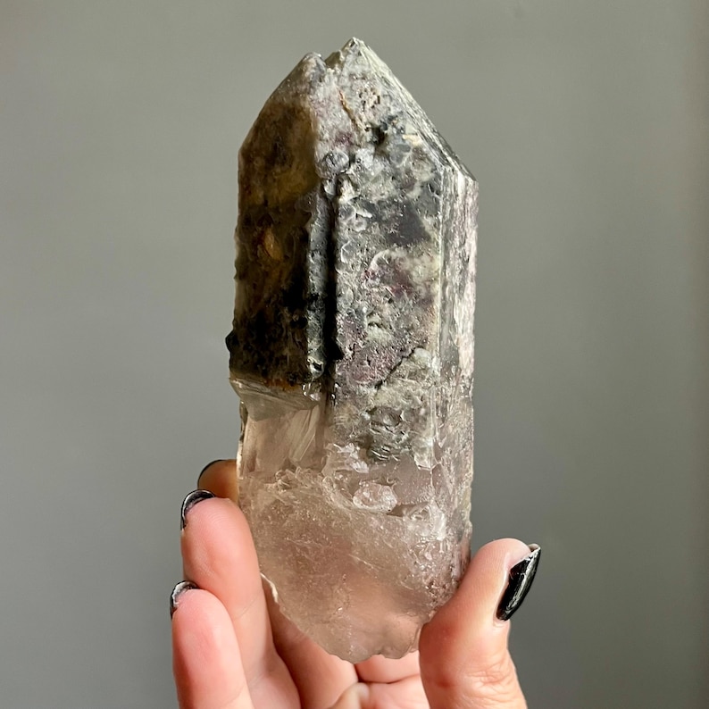 Op de afbeelding: Een natuurlijke kwartskristal met een puntige punt. Het kristal heeft een mix van kleuren, waaronder helder, grijs en paars. Het kristal wordt in een hand gehouden.