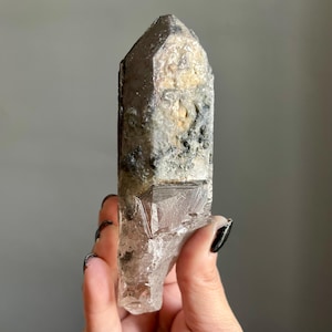Op de afbeelding: Een natuurlijke kwartskristal met een rokerige, doorschijnende uitstraling. De kristal heeft een puntige punt en een ruw, getextureerd oppervlak met grijze, zwarte en beige kleurgebieden. De kristal wordt tegen een grijze achtergrond gehouden.