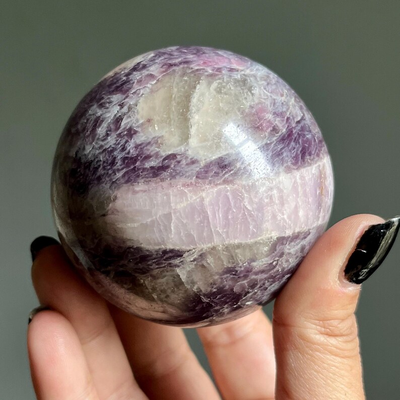 May include: A polished spherical stone featuring a marbled pattern of deep purple, lavender, and translucent white mineral inclusions. The surface is smooth and reflective, showcasing natural crystalline textures and varied color bands throughout.