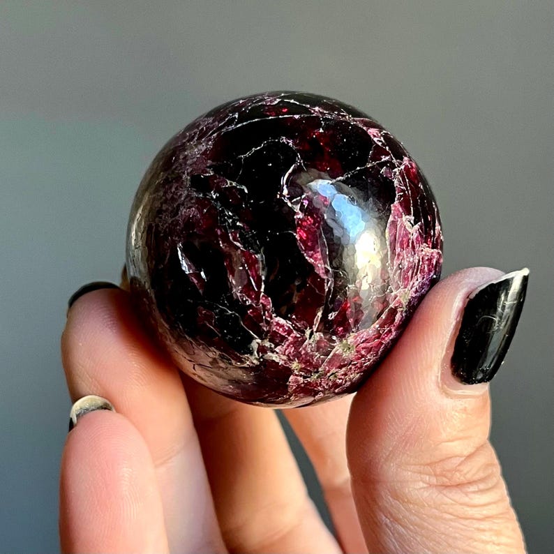 May include: A polished, spherical gemstone with a deep burgundy color and intricate veining. The surface reflects light, showcasing a mix of dark and lighter red hues. The gemstone is held in a hand, highlighting its size and shape.