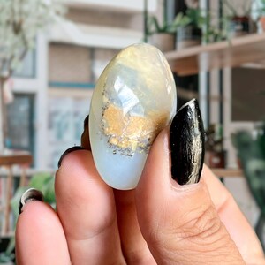 Op de afbeelding: Een gladde, ovale, witte opaal edelsteen met gouden en zilveren insluitsels. De edelsteen wordt in een hand gehouden.