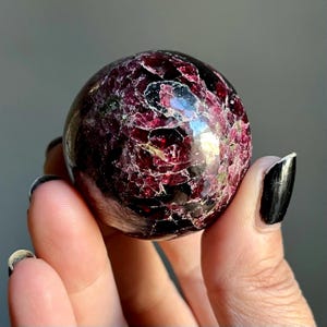 May include: A polished, spherical gemstone with a deep burgundy color and a cracked, textured surface. The sphere is held in a hand, showcasing its intricate patterns and natural beauty. This decorative sphere is a unique addition to any collection.