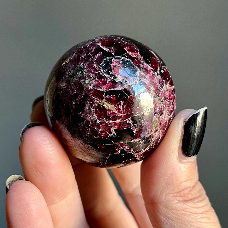 May include: A polished, spherical gemstone with a deep burgundy color and a cracked, textured surface. The sphere is held in a hand, showcasing its intricate patterns and natural beauty. This decorative sphere is a unique addition to any collection.