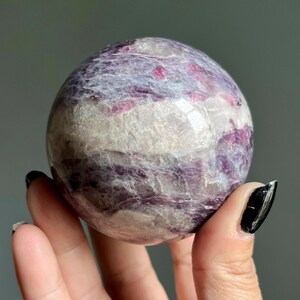 May include: A polished spherical stone featuring a marbled pattern of deep purple, magenta, and white mineral inclusions. The surface is smooth and reflective, showcasing natural crystalline textures and varied color bands throughout the sphere.