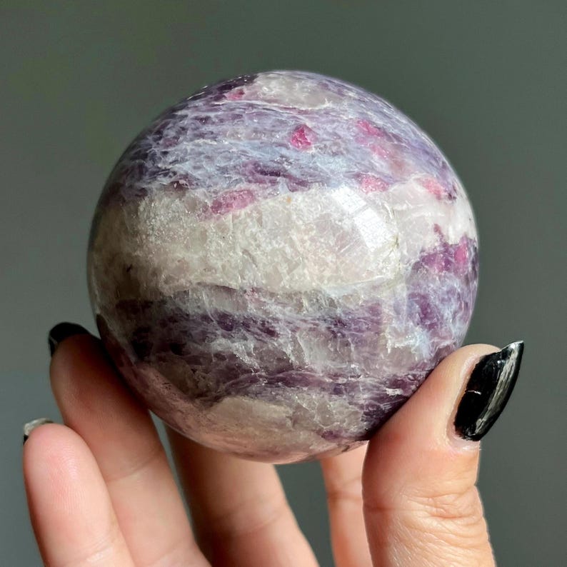 May include: A polished spherical stone featuring a marbled pattern of deep purple, magenta, and white mineral inclusions. The surface is smooth and reflective, showcasing natural crystalline textures and varied color bands throughout the sphere.