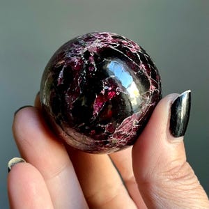 May include: A polished, spherical gemstone with a deep burgundy and black color scheme. The surface of the sphere has a glossy finish with visible internal patterns and inclusions. The gemstone is held in a hand.