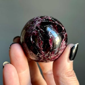 May include: A polished, spherical gemstone with a deep burgundy color and intricate veining. The surface reflects light, showcasing a mix of dark red and lighter pinkish-red hues. The gemstone is held in a hand, highlighting its size and shape.