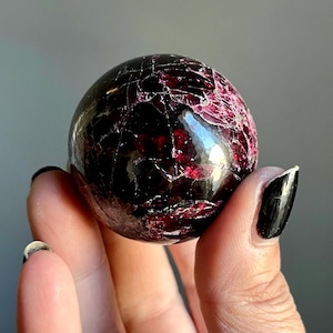 May include: A polished, spherical gemstone with a deep burgundy color and a network of fine cracks. The surface reflects light, highlighting the rich hues and natural patterns of the stone. The gemstone is held in a hand.