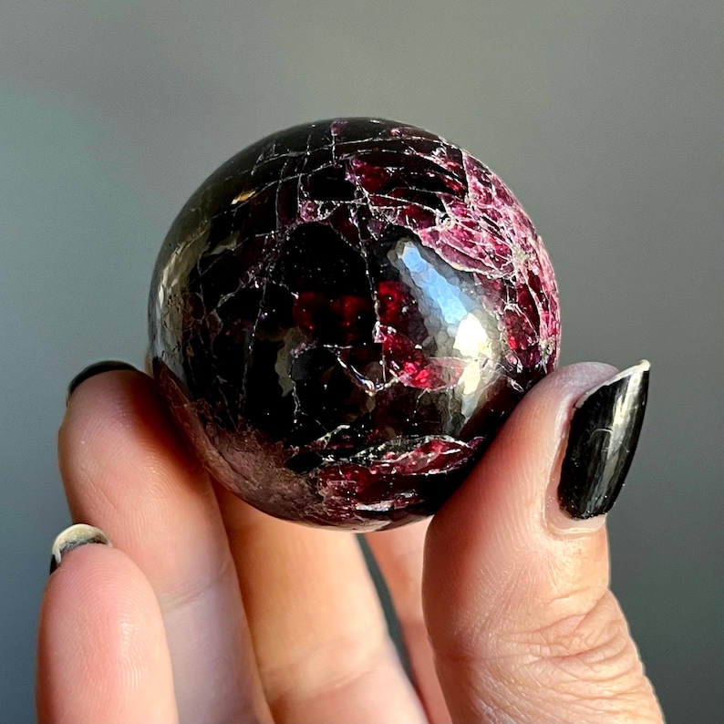 May include: A polished, spherical gemstone with a deep burgundy color and a network of fine cracks. The surface reflects light, highlighting the rich hues and natural patterns of the stone. The gemstone is held in a hand.