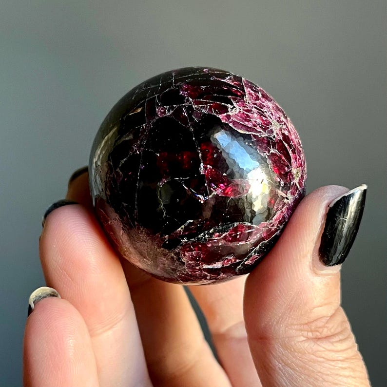 May include: A polished, spherical gemstone with a deep burgundy color and a network of fine cracks. The surface reflects light, highlighting the stone's texture and internal patterns. The gemstone is held in a hand.