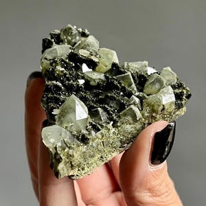 May include: A mineral specimen featuring a cluster of translucent, pale green, pointed crystals embedded within a dark, metallic, textured matrix. The crystalline structure displays sharp, geometric facets and a natural, raw appearance.