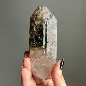 Op de afbeelding: Een natuurlijk kwartskristalpunt met een ruw, getextureerd oppervlak. Het kristal heeft een kleurengradi&euml;nt, waaronder heldere, rokerige en zwarte gebieden. Het kristal wordt in een hand gehouden tegen een grijze achtergrond.