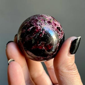 May include: A polished, spherical gemstone with a deep burgundy base and a network of lighter red and green veins across its surface. The sphere is held in a hand, showcasing its reflective, glossy finish. The gemstone appears to be a decorative item.