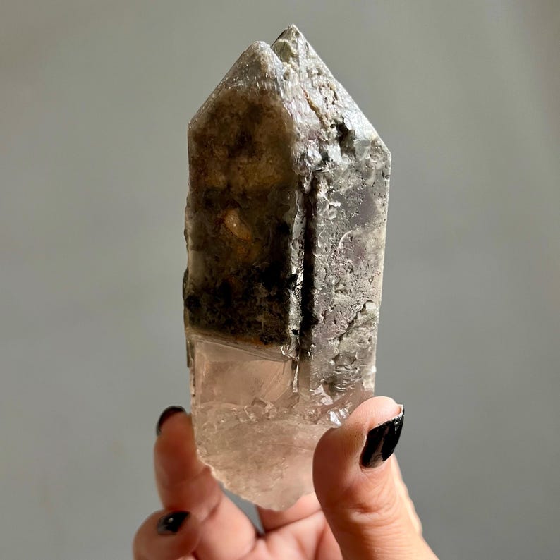 Op de afbeelding: Een natuurlijk kristalpunt met een ruw, getextureerd oppervlak. Het kristal heeft een kleurovergang, van een helder, lichtroze aan de basis naar een rokerig grijs en bruin aan de top. Het kristal wordt in een hand gehouden.
