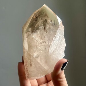 May include: A clear quartz crystal with a pointed top and a rough, textured side. The crystal has a light brown hue and is held in a hand against a gray background. The crystal is a natural, raw gemstone.